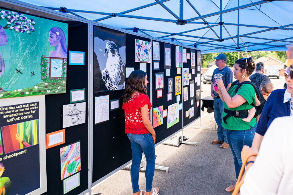People viewing a public art display with children’s drawings and paintings under a canopy tent.