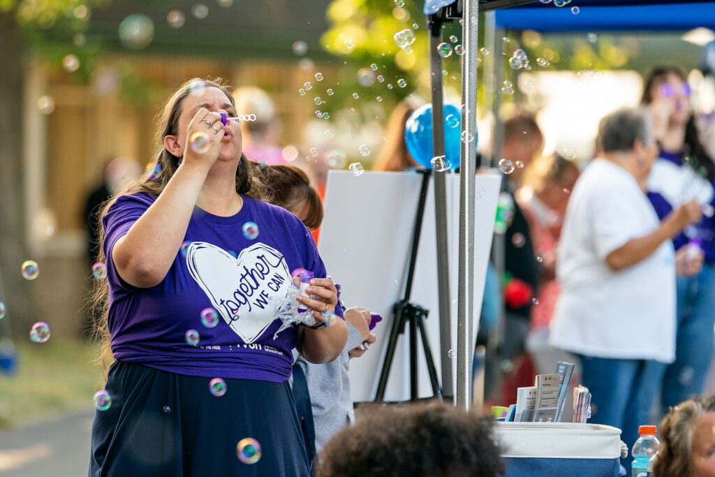 Woman in a purple “Together We Can” shirt blowing bubbles at an outdoor community event.