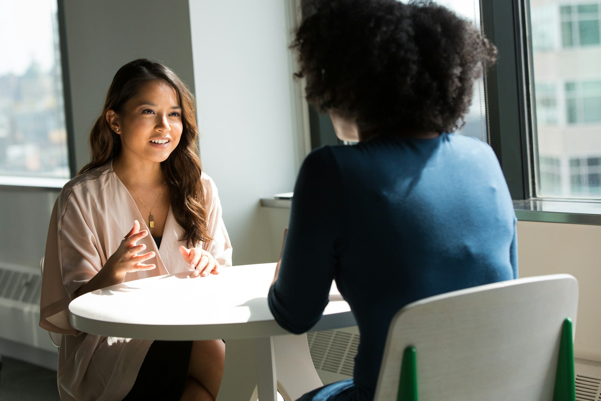 Two women talking across a table in a sunlit office, with one gesturing as she speaks during a private meeting.