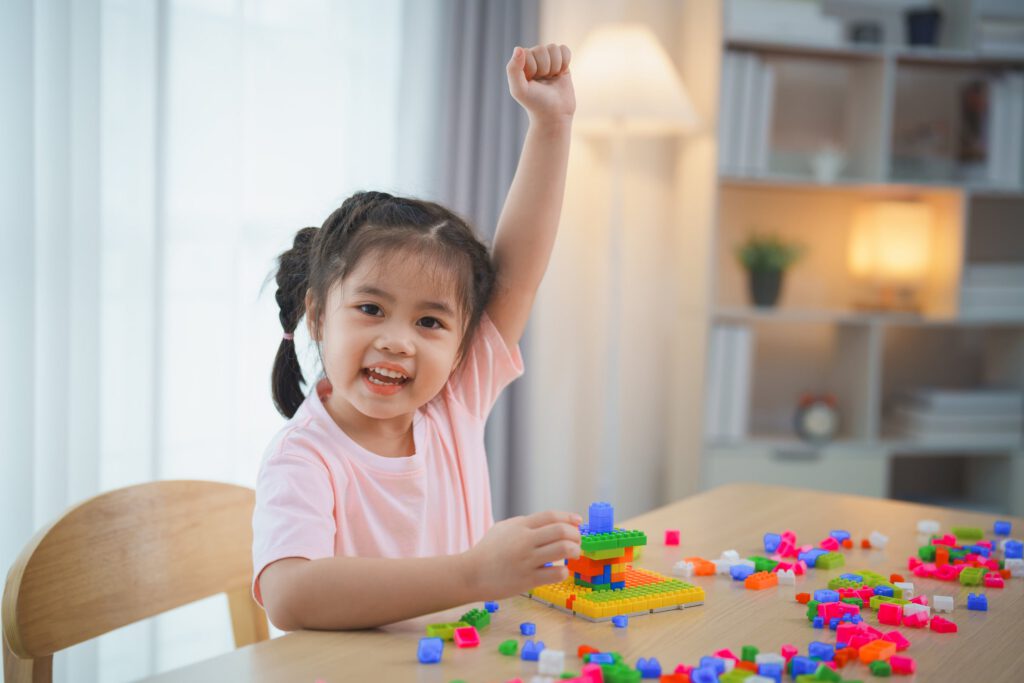 Smiling child building blocks and raising hand at a table